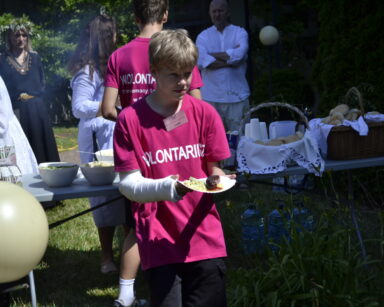 Zbliżenie. Młody wolontariusz z ręką w gipsie niesie talerzyk z grillowaną kiełbasą. W tle grupa ludzi przy grillu.