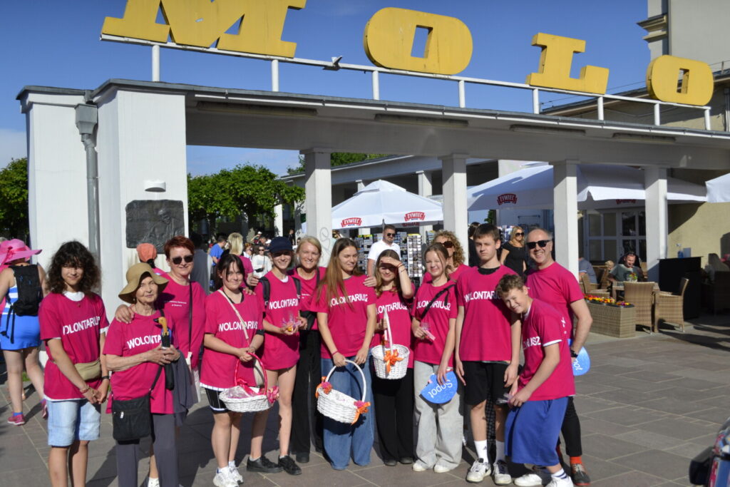 Brama. Napis "Molo". Pod nim grupa wolontariuszy w różowych koszulkach. W rękach trzymają kolorowe kosze. Pozują do zdjęcia.