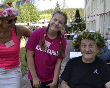 Ogród. Dwie wolontariuszki pozują do zdjęcia z uśmiechniętym seniorem. Senior na głowie ma wianek z żywych kwiatów.