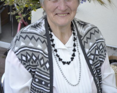 Zbliżenie. Seniorka w białej koszuli, czarno srebrnych koralach i wianku na głowie pozuje do zdjęcia. Kobieta uśmiecha się.