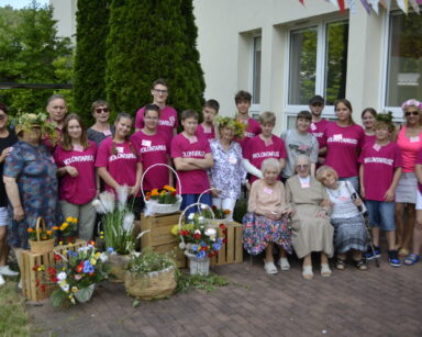 Ogród. Grupa wolontariuszy w różnym wieku pozuje do wspólnego zdjęcia. Obok w koszach, kolorowe kwiaty.