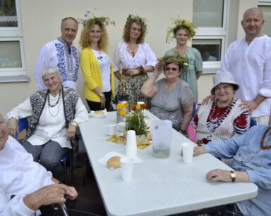 Zbliżenie. Pięć osób z DDP i DPS stoi przy stole gdzie siedzą seniorzy. Wszyscy pozują do zdjęcia. 4 kobiety maja wianki.