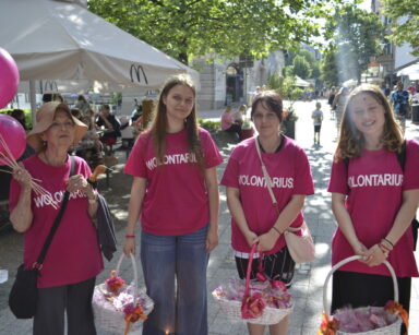 Miasto, promenada. Cztery wolontariuszki w różowych koszulkach, z napisem Wolontariat i koszami w dłoni pozują do zdjęcia.