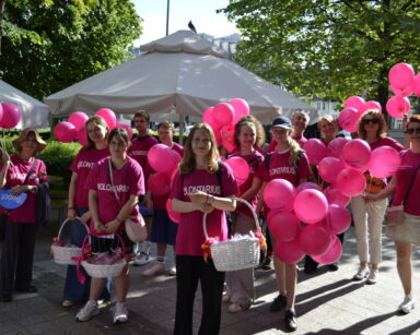Miasto, promenada, drzewa, parasol. Grupa wolontariuszy w różowych koszulkach z koszami i balonami pozuje do zdjęcia.