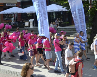 Miasto, promenada, drzewa, parasole. Maszeruje grupa ludzi w różowych koszulkach oraz klub pływacki Sopot.