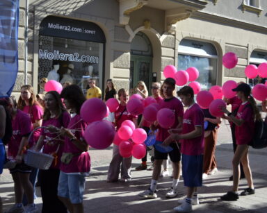 Miasto, promenada, kamienice, sklepy. Maszerują wolontariusze w różowych koszulkach i z balonami w rękach .