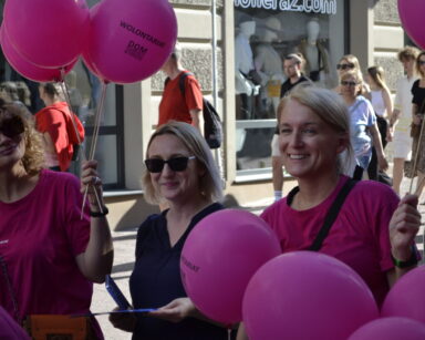 Promenada, kamienice, sklepy. Zbliżenie na trzy kobiety dwie trzymają balony z napisem Wolontariat. W tle grupa ludzi.