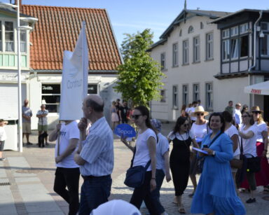 Promenada, kamieniczki, parasole. Grupa ludzi maszeruje, kobiety w rękach trzymają wachlarze z napisem Sopot.