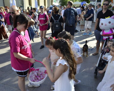 Miasto, promenada, tłum ludzi. Czworo dzieci w jasnych ubrankach podeszło do wolontariuszki po słodkości z koszyka.
