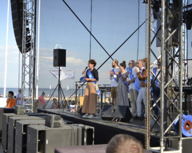 Scena, widok na plażę i morze. Na scenie pani Prezydentka Sopotu z mikrofonem w ręku i grupka osób klaszczących.