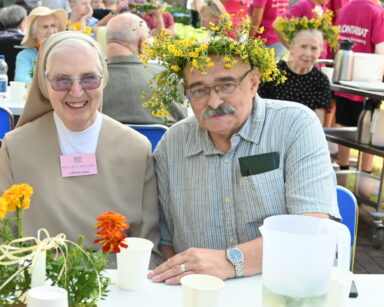Zbliżenie. Senior w wianku na głowie i koszuli w kratę siedzi koło wolontariuszki, siostry zakonnej. Na stole kwiaty, kubki.