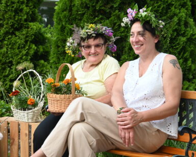 Zbliżenie. Młoda kobieta i seniorka w wiankach na głowie siedzą na ławeczce. Seniorka trzyma kosz z kwiatami.