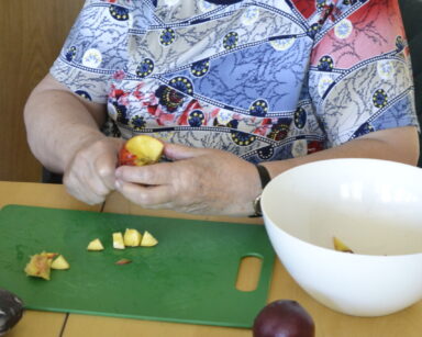 Zbliżenie. Seniorka kroi w kostkę jabłko. Obok na stole stoi biała miska i leży kilka brzoskwiń.