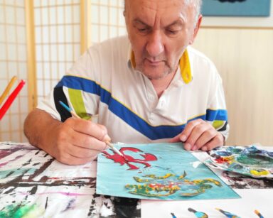 Zbliżanie. Senior siedzi przy stole. W ręku trzyma pędzel, obok farby. Senior maluje czerwonego kraba na niebieskim tle.