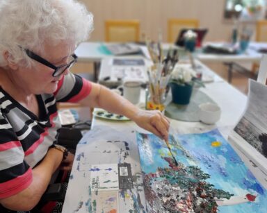 Zbliżenie. Seniorka lewą ręką maluje farbami obraz. Na nim latarnia morska, fale, skała, niebo i słońce.