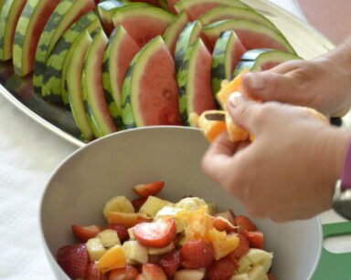 Zbliżenie. Na tacy leżą kawałki pokrojonego arbuza. Obok w białej misce pokrojone truskawki, banany i pomarańcze.