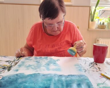 Zbliżanie. Seniora siedzi przy stole. W ręku trzyma stempel z farbą. Przed seniorką leży kartka pomalowana na niebiesko.