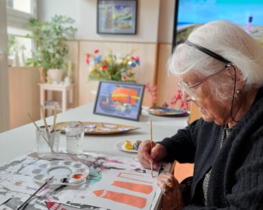 Zbliżenie. Seniorka maluje latarnię morską. Obok leżą przykładowe wycięte obrazki latarni morskich. Na stole farby, pędzle.