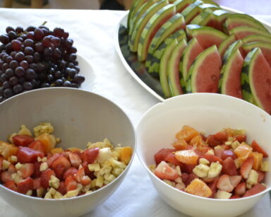 Zbliżenie. Na stole w misce leżą pokrojone owoce, obok na talerzyku leżą ciemne winogrona oraz pokrojony w kawałki arbuz.