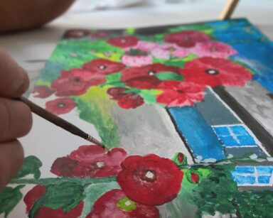 Zbliżenie. Stół. Ręka trzyma pędzel z farbą. Na stole obraz a na nim czerwone malwy, niebieskie niebo, szary domek.