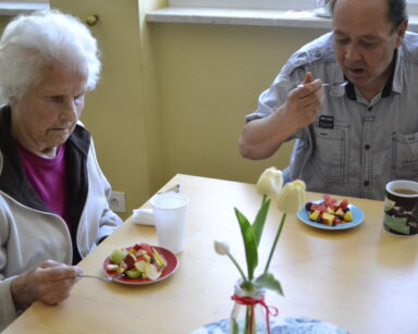 Zbliżenie. Para seniorów siedzi przy stole i je sałatkę owocową. Obok stoją kubeczki. Pośrodku stołu stoi wazon z kwiatami.