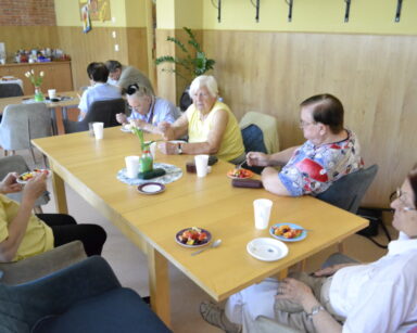 Sala. Grupa seniorów siedzi przy dwóch stołach i je z talerzyków owocowe sałatki. W tle na blacie kuchennym stoją naczynia.