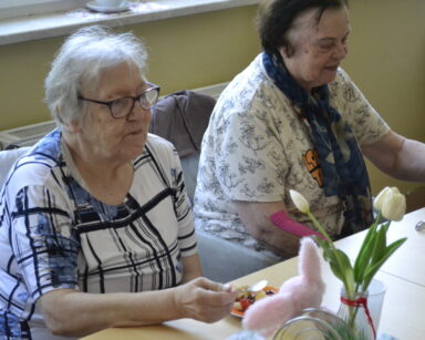 Zbliżenie. Dwie seniorki siedzą przy stole i jedzą owocowe sałatki. Na stole stoi wazon z białym tulipanem oraz zajączek.