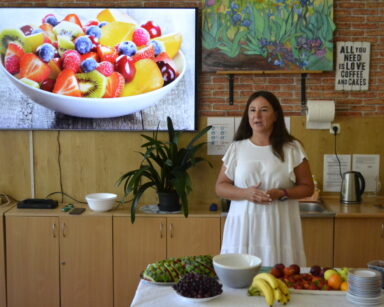 Sala. Kobieta w białej sukience stoi przy stole, na którym stoją owoce. W tle na monitorze, zdjęcie sałatki owocowej.