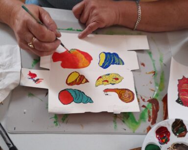 Zbliżenie. Dłonie seniorki. Kobieta maluje na stole obrazek farbami. Różne rodzaje muszli morskich. W ręku trzyma pędzel.