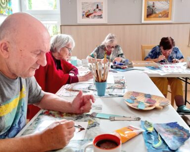 Sala. Przy stole seniorzy malują obrazy. Na stole leżą wycięte elementy obrazków jak fale morskie, latarnie morskie.