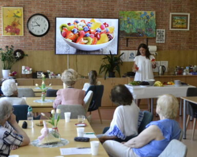 Sala. Seniorzy siedzą przy stolikach. Obok kobieta stoi przy stole. W tle na monitorze, zdjęcie sałatki owocowej.