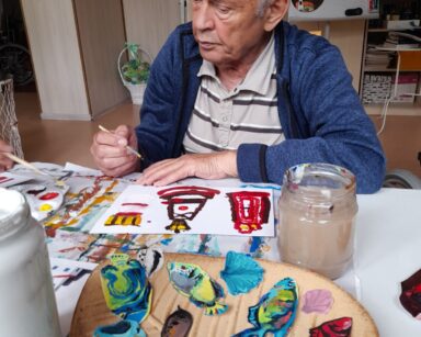 Zbliżenie. Senior siedzi przy stole. Maluje latarnie morskie. Przed nim wycięta z kartki kolorowa fauna i flora morska.