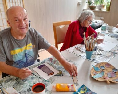Zbliżenie. Dwóch seniorów przy stole maluje farbami. Na rysunkach są latarnie morskie obok stoją na stole farby, pędzle.