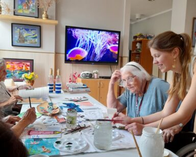 Zbliżenie. Seniorzy siedzą przy stole i malują obrazy. Obok seniorki pochyla się młoda kobieta i maluje pędzlem.
