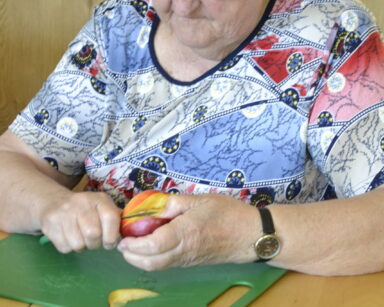 Zbliżenie. Seniorka w kolorowej bluzce kroi w ręku brzoskwinię. Obok na stole deska do krojenia i kilka brzoskwiń.