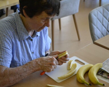 Zbliżenie. Seniorka w koszuli w kratkę kroi w rękach banana. Na desce leżą kawałki owocu, obok trzy banany w całości.