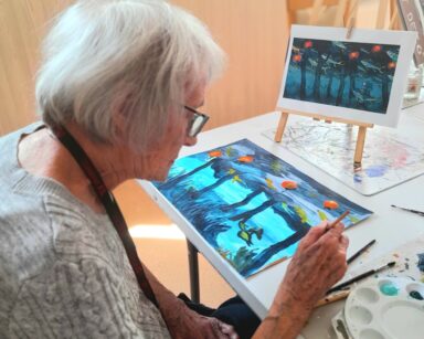 Zbliżenie. Seniorka siedzi przy stole, maluje obraz. Obok paleta z farbami. Na stole sztaluga ze zdjęciem.