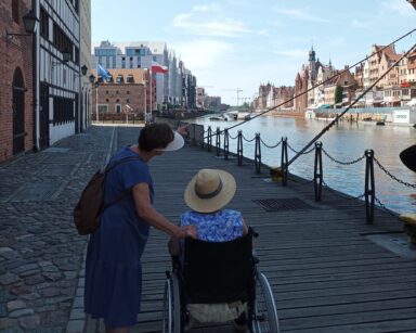 Drewniany podest, kamienice, seniorka na wózku obok kobieta wskazuje na rzekę. W tle kamienice starego miasta Gdańsk.