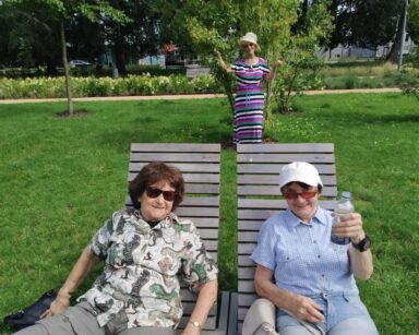 Ogród. Dwie seniorki pozują do zdjęcia siedząc na drewnianych siedziskach. W tle seniorka w kapeluszu stoi przy drzewie.