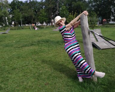 Park. Seniorka w sukience w paski trzyma rękoma drewniany palik od hamaka. Seniorka pozuje do zdjęcia.