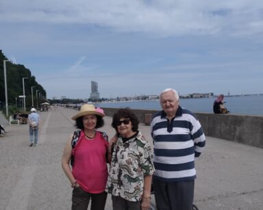 Deptak, falochron, morze. Na deptaku grupa ludzi, trzech seniorów pozuje do wspólnego zdjęcia. W tle Sea Towers.