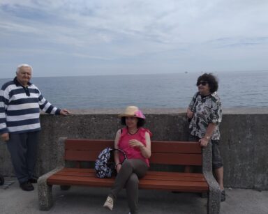 Ławka, falochron, morze. Trzech seniorów pozuje do zdjęcia. Jedna seniorka siedzi na ławce, obok stoi dwóch seniorów.