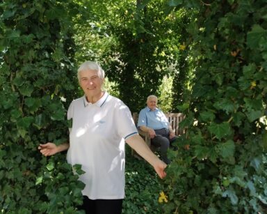 Zbliżenie. Ogród, drzewa pokryte bluszczem. Między drzewami stoi seniorka, w tle na ławeczce siedzi senior.