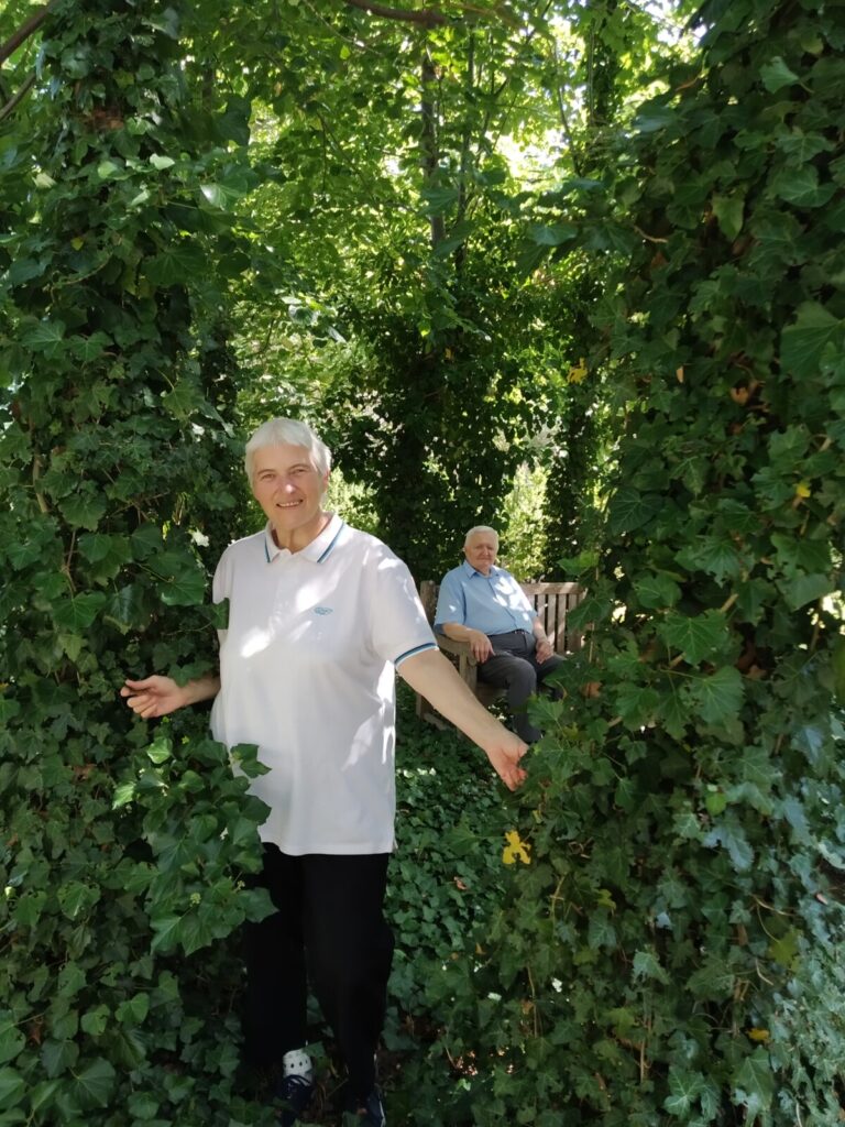 Zbliżenie. Ogród, drzewa pokryte bluszczem. Między drzewami stoi seniorka, w tle na ławeczce siedzi senior.