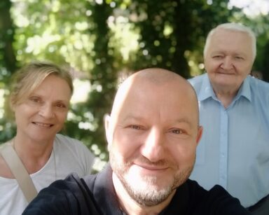 Zbliżenie. Trzy osoby pozują do selfie. Mężczyzna w ciemnym polo, kobieta w jasnej bluzce i senior niebieskiej koszuli.