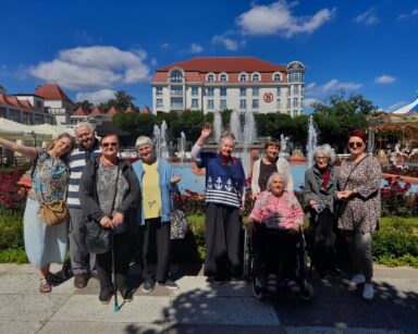 Grupa osób pozuje do wspólnego zdjęcia. W tle fontanna, karuzela, hotel oraz parasole.