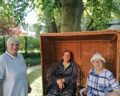 Zbliżenie. Trzy uśmiechnięte seniorki pozują do zdjęcia. Dwie siedzą w plażowym koszu, jedna stoi obok. W tle drzewo.