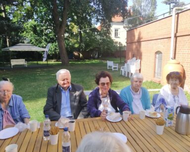 Zbliżenie. Ogród. Przy stole siedzą seniorzy, na stole woda i przekąski. W tle rozkładana altana, ławka, białe krzesła.