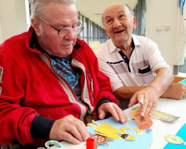 Sala. Dwóch uśmiechniętych seniorów wykleja kolaż. Na obrazie rybak, ster. Obok senior seniorowi, podaje element wyklejanki.