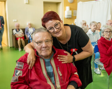 Zbliżenie. Kobieta w czarnej sukience z patriotycznym kotylionem, przytula seniora siedzącego na wózku inwalidzkim.